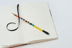A colorblock Blackwing pencil featuring Mickey Mouse faces rests on an open blank notebook with a black ribbon bookmark, shot on a clean white background.