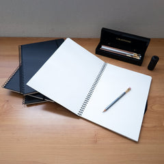 a stack of Blackwing Spiral Notebooks on a wood desk that also features a 602 pencil a one-step sharpener and a piano box of blackwing pencils