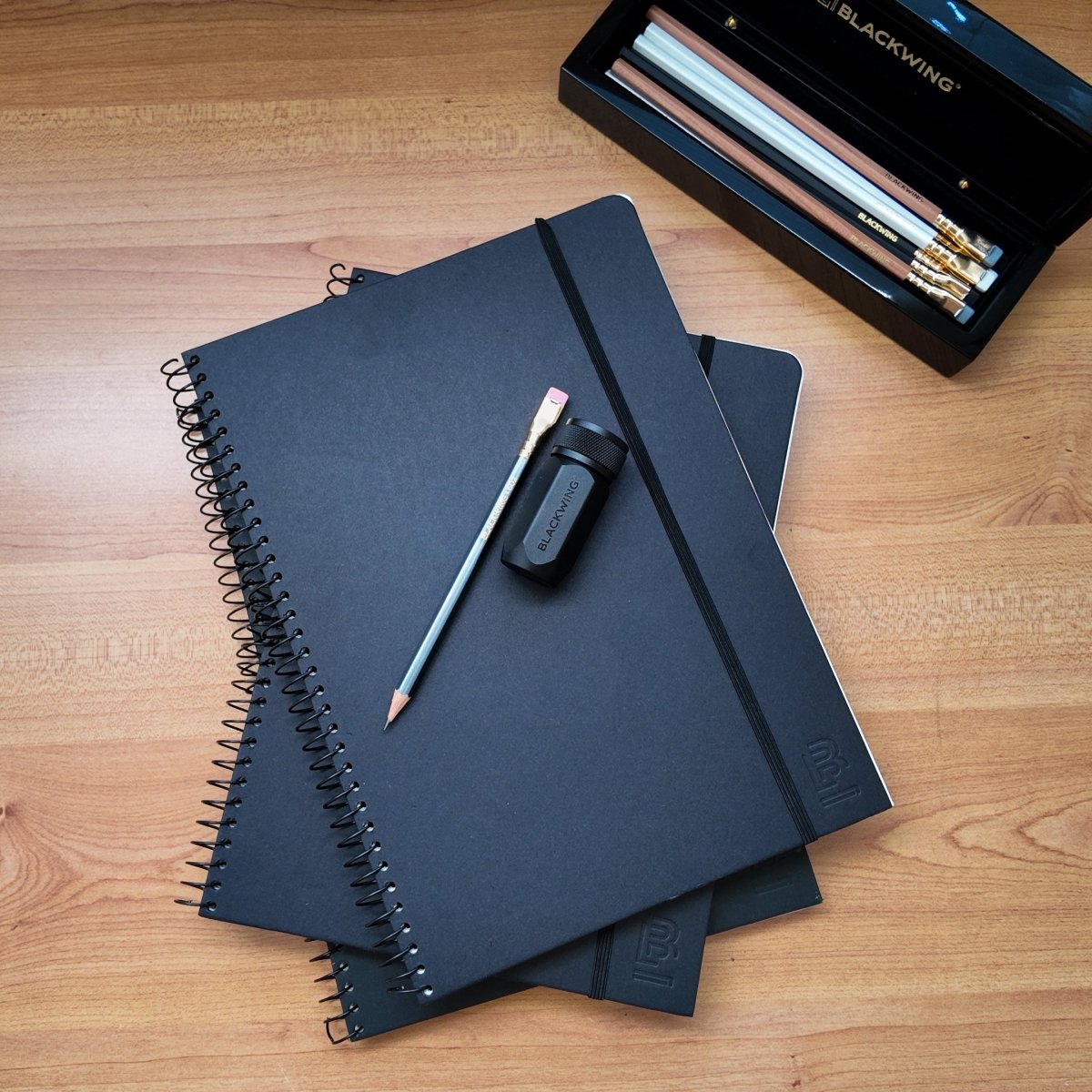 Stack of 3 Blackwing Spiral Notebooks on a wooden desktop with a 602 pencil, a one-step sharpener and a piano box of Blackwing pencils