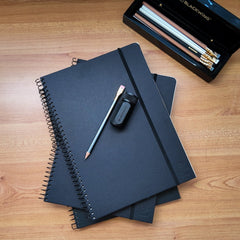 Stack of 3 Blackwing Spiral Notebooks on a wooden desktop with a 602 pencil, a one-step sharpener and a piano box of Blackwing pencils