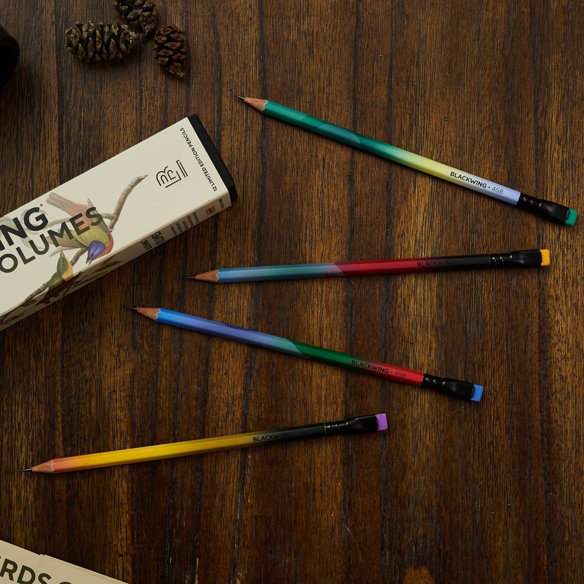 4 gradient pencils with black ferrule and colorful erasers on top of a dark wood desk next to pinecones and a Blackwing Volumes box featuring birds