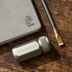 Blackwing One-Step Long Point Sharpener in grey, next to a grey pencil and grey slate notebook